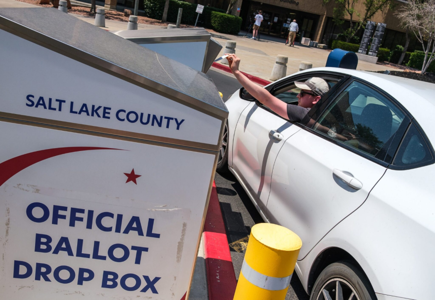 A person leans out of their car in a parking lot to drop a ballot into a drop box, labeled Salt Lake County Official Ballot Drop Box.