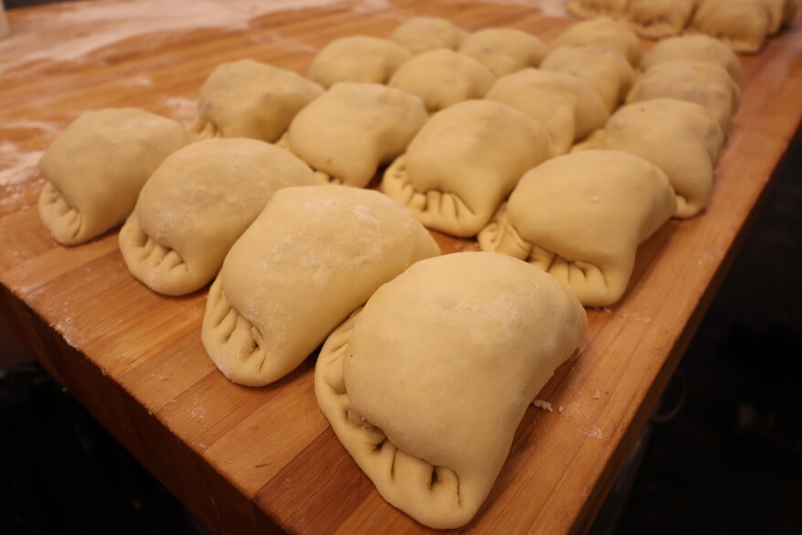 A 4x4 row of pasties sits on a large wooden table. The half-moon shaped pastries are crimped along the outer edge. The dough is still a light cream color. After they're baked, they'll be a golden brown!