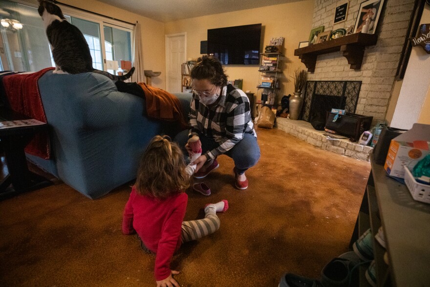 A woman helps a young girl put on her shoes in a living room. 