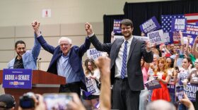 Three men stand holding hands in the air in celebration in front of a crowd of people.