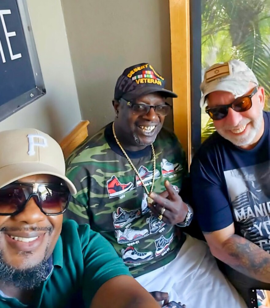 Selfie shows three men wearing baseball caps, sitting and smiling into the camera