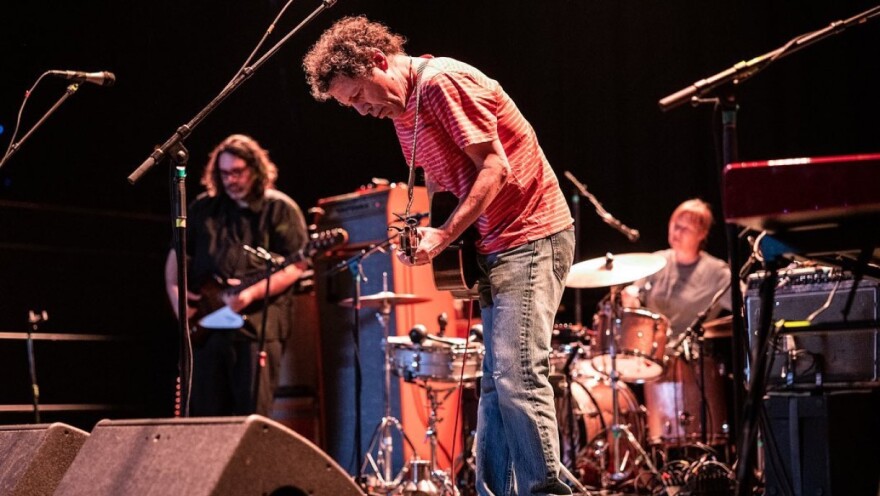 A man on stage plays a guitar while looking at the ground, with a bass player and a drummer out of focus in the background.