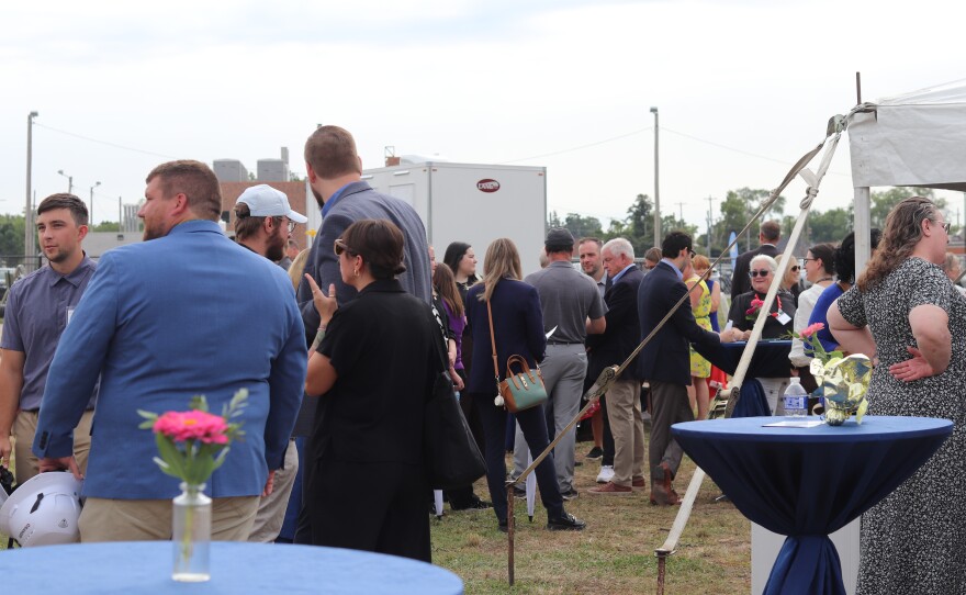 A crowd of people gathers outdoors at an event, standing and talking near cocktail tables with flowers, under a cloudy sky.