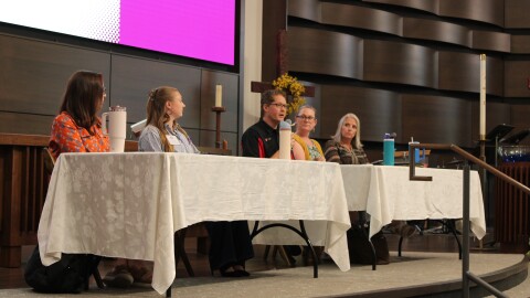 A photo of five adults sit at two tables on top of a stage.