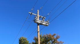 A upward looking view of a powerline and pole. Attached to the top is a horizontal metal box that is the "smart switch." The tops of some green leaves and trees are visible at the bottom of the image. 