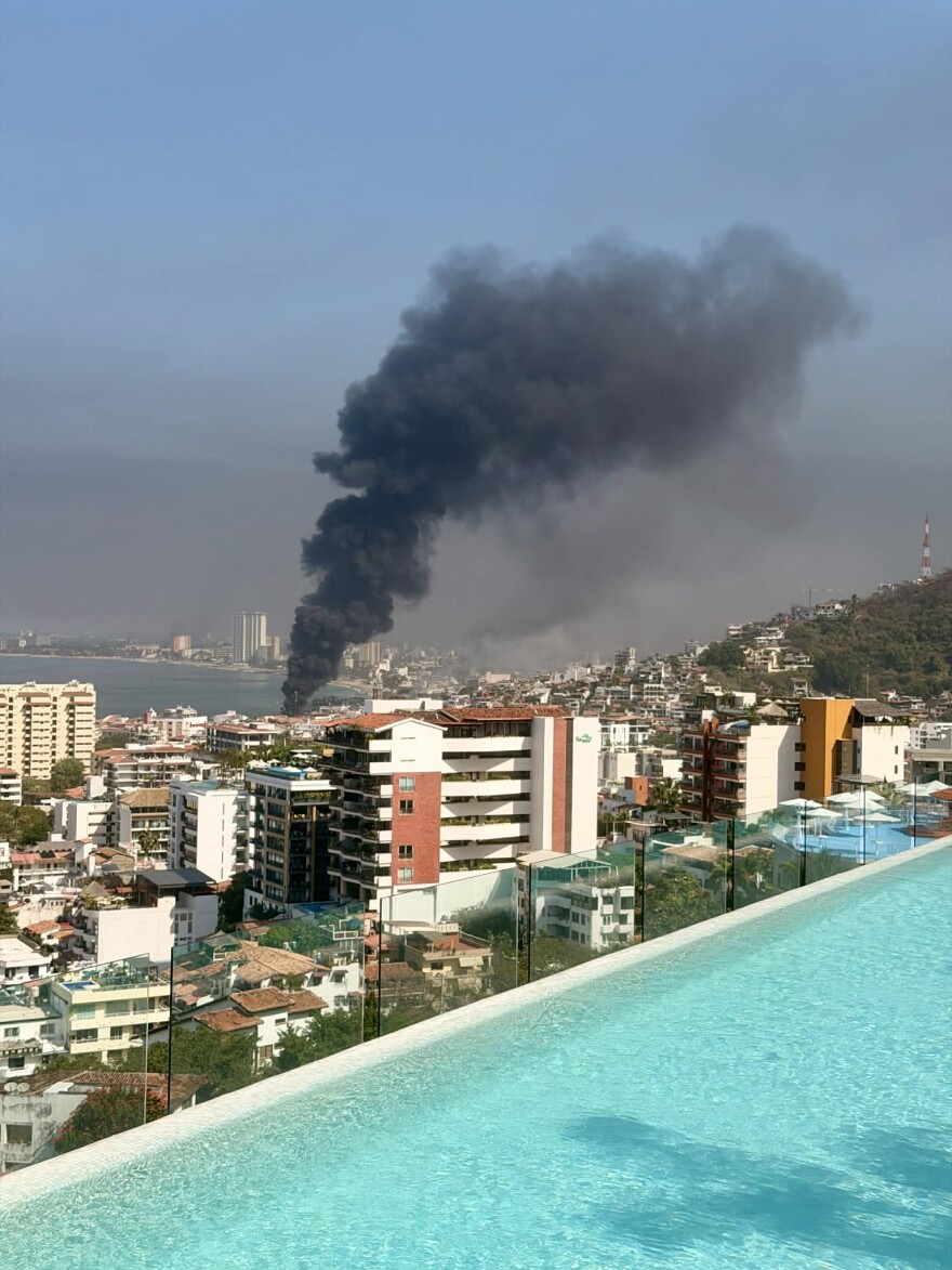 The edge of a rooftop pool in the foreground, a dark grey cloud of smoke from an unknown building billows over the white and orange buildings in Puerto Vallarta, Mexico. 