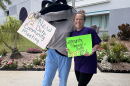 A person inside of a blue and gray stingray mascot costume stands holding a white sign, while the other arm is around the shoulder of a woman wearing a purple shirt and black leggings, holding a green sign. Both signs note a community meeting taking place on Oct. 12.