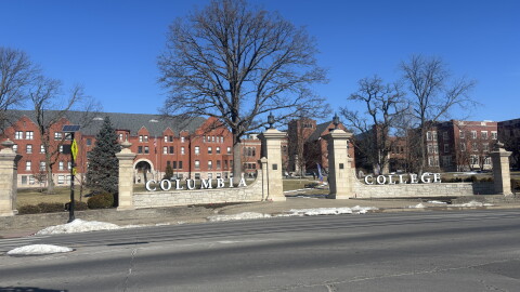Patches of snow cover the ground in front of the gates of Columbia College on a sunny winter day. The word "Columbia" sits on the left gate and "College" on the right with a sidewalk in the middle. 