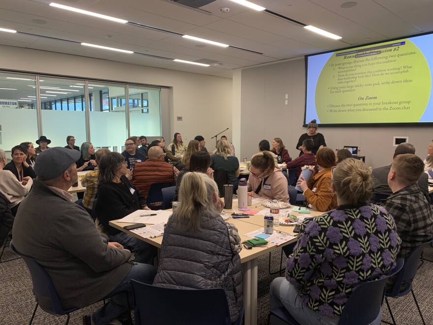 About thirty people sit at tables in the community room at the Logan Library, listening to Maureen Boyle as she presents at the front. The current slide asks attendees to have round table discussions about their hopes for the coalition and how it will work.