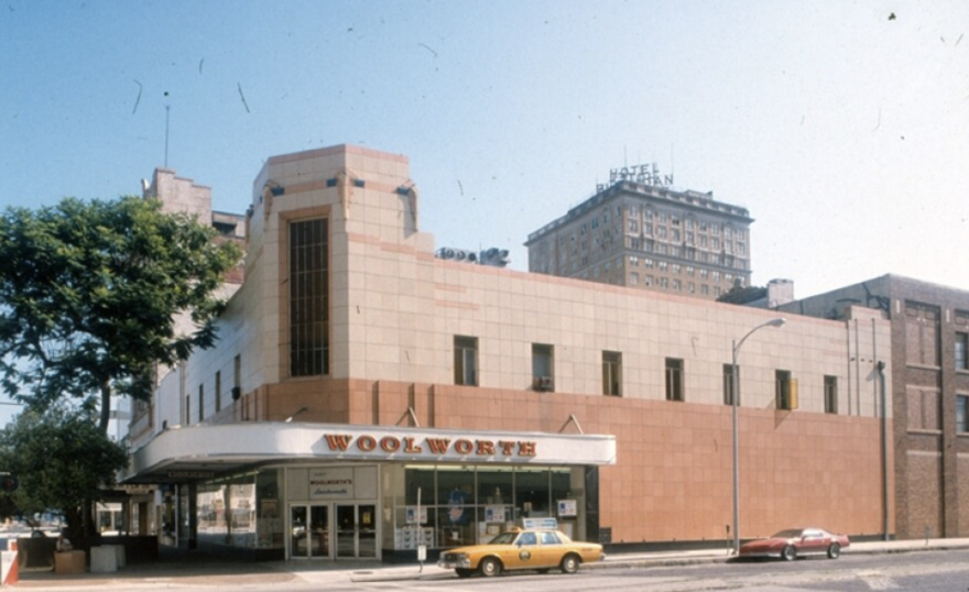 An old, tan colored building on a street corner with dark orange letters spelling WOOLWORTH
