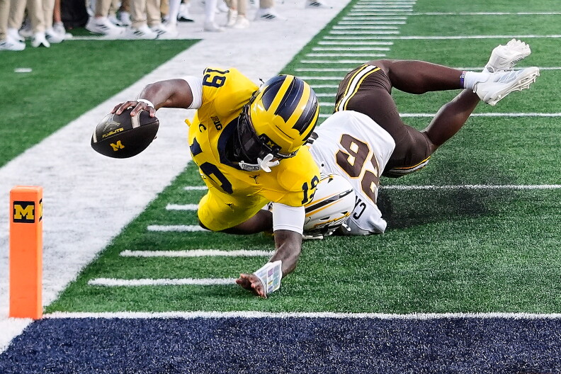 Michigan Wolverines football player dives toward the end zone with the ball as a defender tackles him near the orange pylon.