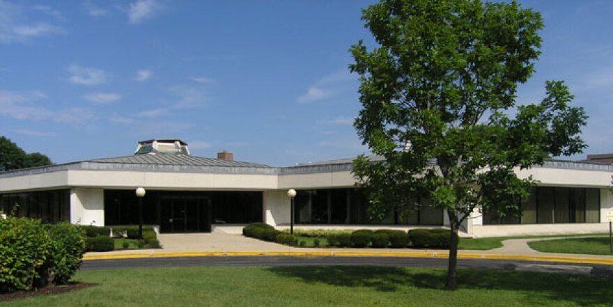 A one-story white building is pictured, with a blue sky, green lawn and a tree in front.