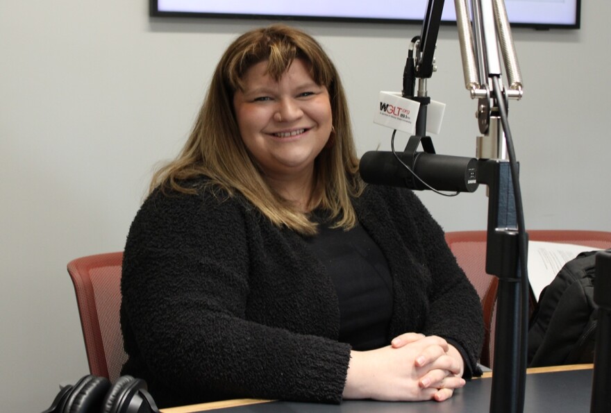 A woman dressed in black sits with her hands folded at a desk, smiling toward the camera. A microphone is in front of her and headphones to her side. 