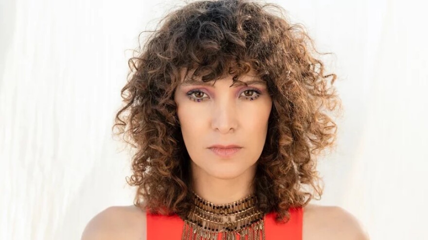 A young woman with curly brown hair looks intensely at the camera with a light background behind her.