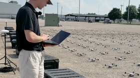 A man holds a laptop in front of a gravel lot filled with drones