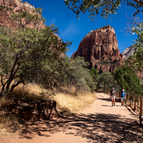 Two people walk along a trail with beautiful red rock formations in the background.