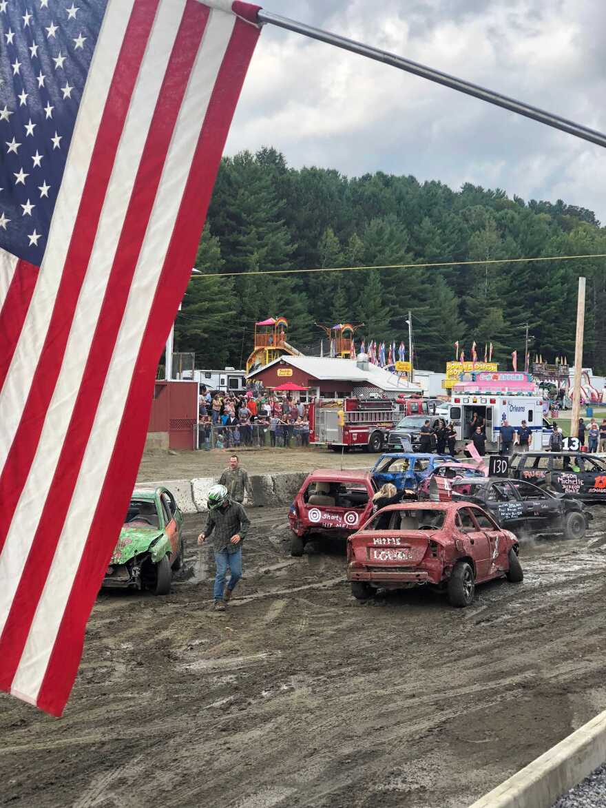 The demolition derby at the Orleans County Fair, with mud in the forefront, under an American Flag.