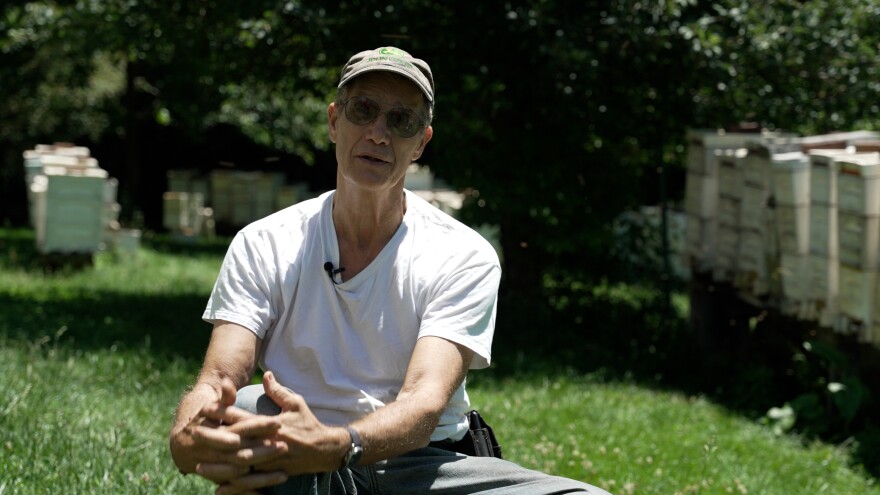 Beekeeper Joe May sitting near his hives. He's wearing a baseball cap and a white t-shirt.