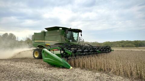 A green combine churns through a tan field of soybeans, spraying chaff out the back