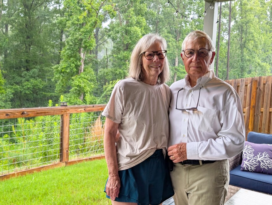 Gail and Rob Sansome stand on their back porch with leafy green trees behind them.