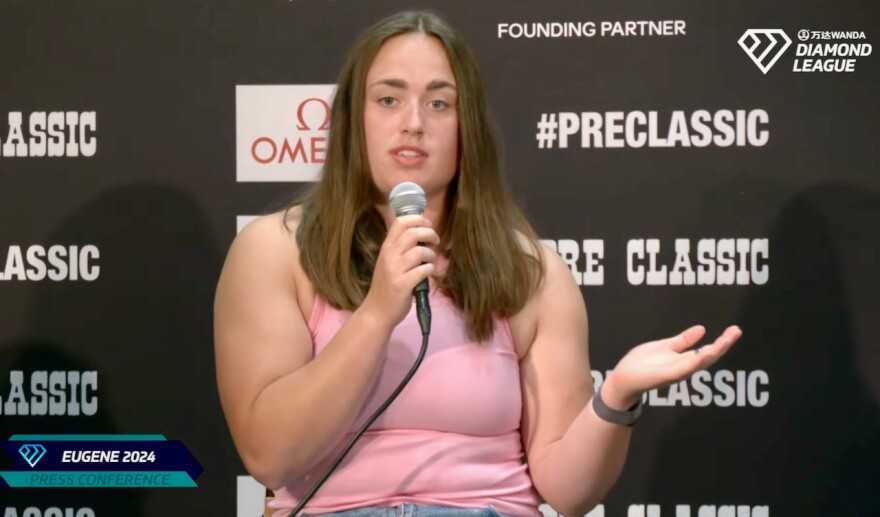 A woman in a pink tank top speaks into a microphone.