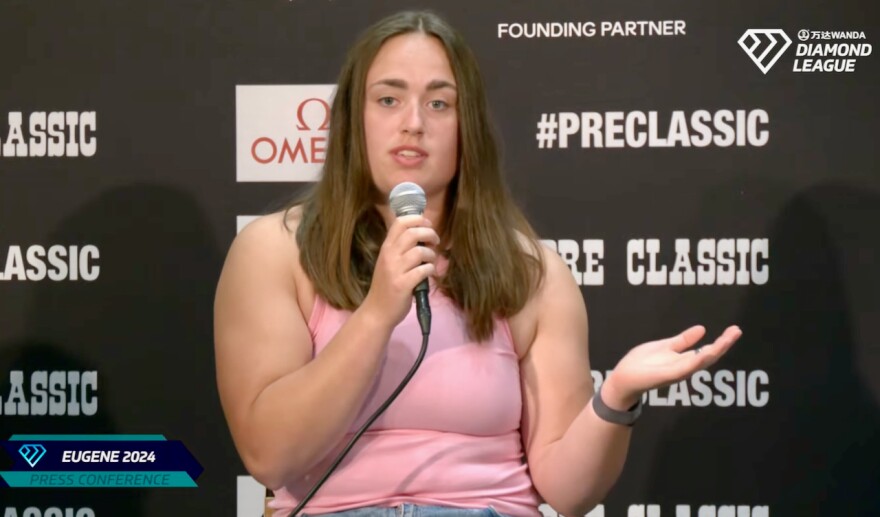 A woman in a pink tank top speaks into a microphone. 