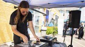 A man in black with long brown hair putting a record onto a records player. He is DJing under a tent in a parking lot. 
