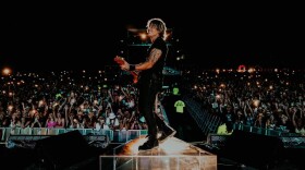 A man smiles while standing on stage and playing electric guitar with a massive crowd of cheering fans in the background.