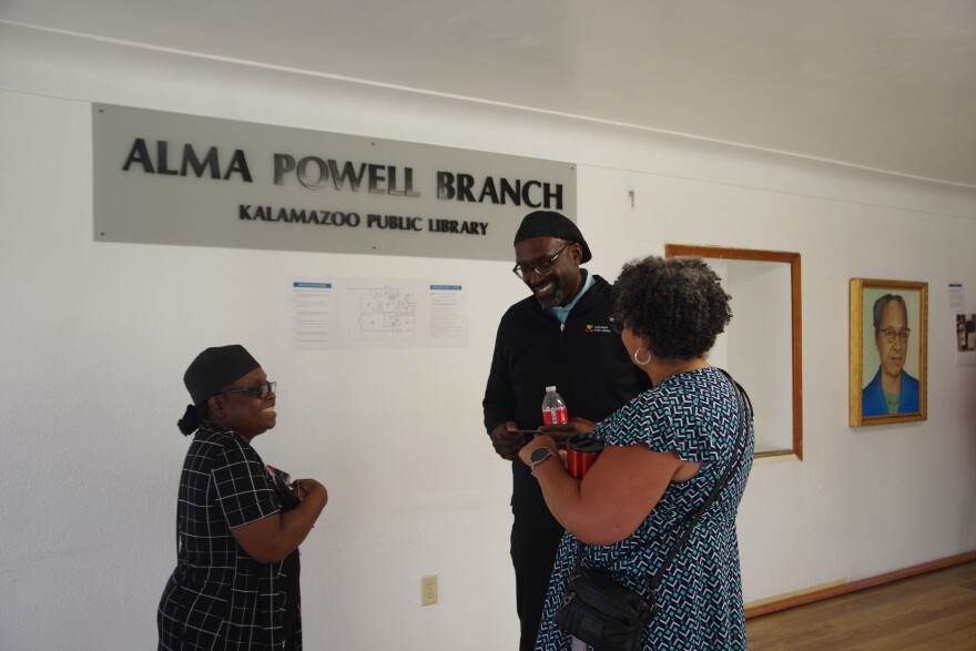 Three people stand in a circle discussing the new temporary Alma Powell Branch location. Two of the attendees can be seen smiling as they converse. A sign reading "Alma Powell Branch, Kalamazoo Public Library" can be seen above them, with a portrait of the branch's name sake sitting on the wall on the right of the image.  