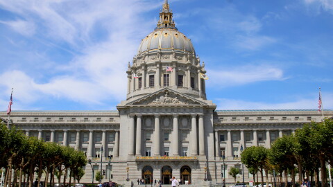A view of the front of city hall with a few trees in the foreground and a blue sky with wispy clouds behind it. The city hall building is long and rectangular with columns in front, gold trim, and a big dome coming out of the center of it. You can see a few people walking in front of the building. 