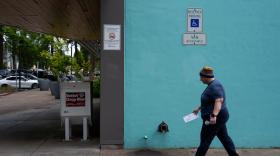 A person carrying an envelope walking on a sidewalk approaching a ballot drop box.