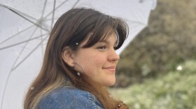 A woman with long brown hair wears a denim jacket and holds a white umbrella. She is smiling and snow is falling. 