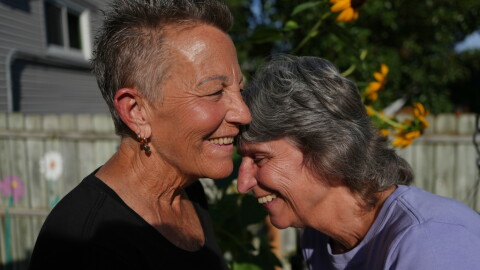 2 older white women laugh in the backyard of a home  , the one on the left has short cropped grayish hair in a black top.... the one on the right has darker hair and a light blue top 