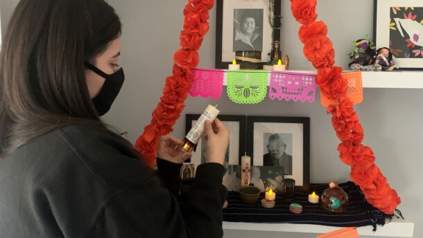 A woman stands in front of a Mexican "ofrenda" with orange paper marigolds and photos of her family. She is holding an unlit candle.