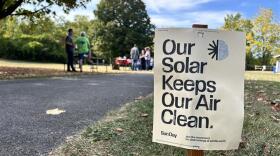 A small sign steaked into the grass along a grassy path, is in the photo's foreground. It reads, "Our Solar Keeps Our Air Clean: Join the movement for clean energy at Sunday.earth." On the left side of the photo frame are a few people surrounding a picnic area, and a white pop up tent in the background.