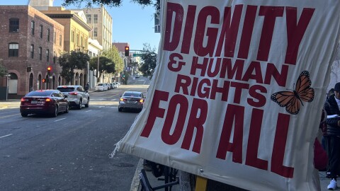 A sign reading "dignity and human rights for all" leans on a tree outside of 630 Sansome Street. 
