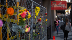 Una valla metálica con cinta de precaución y flores entretejidas. Al fondo, el letrero rojo de Bradleys.