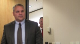 Governor Dunleavy peeks through a door with a man in a suit in the foreground