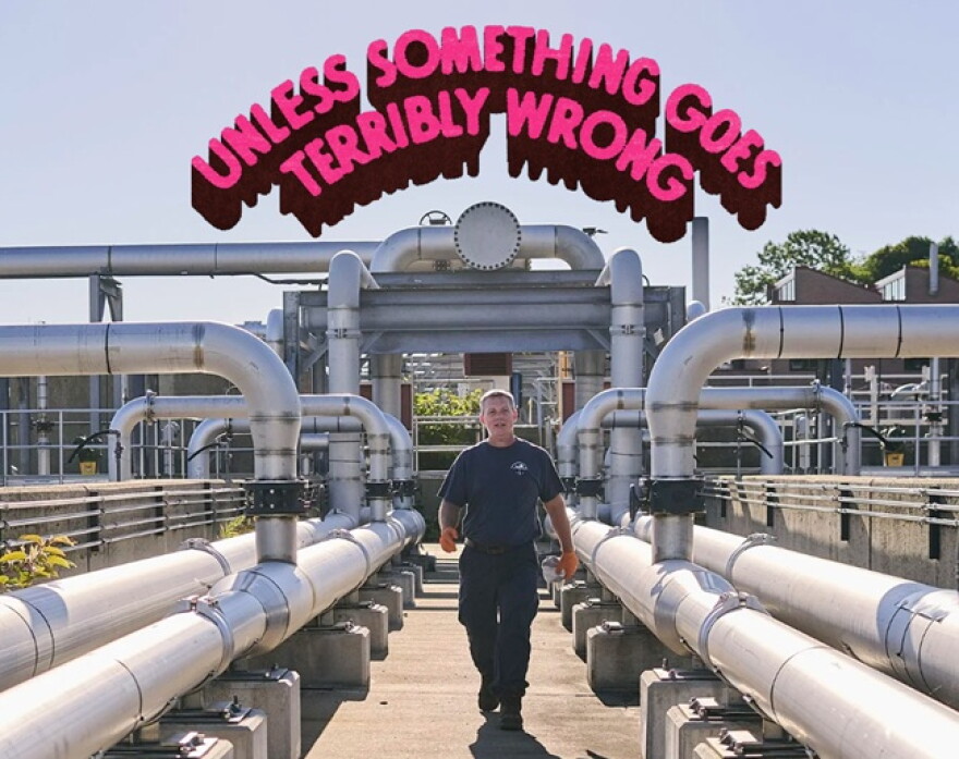 A man walking amid a series of large pipes in a sunny outdoor space; "Unless Something Goes Terribly Wrong" written in a pink arch overhead