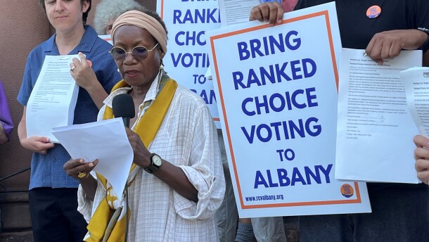 A woman with a loose-fitting button-up shirt and decorative scarf, Ranked Choice Voting Albany Co-Chair Charlotte Collet, speaks into a microphone on the steps in front of Albany City Hall. Supporters behind her hold pages of signatures and signs reading "BRING RANKED CHOICE VOTING TO ALBANY."  