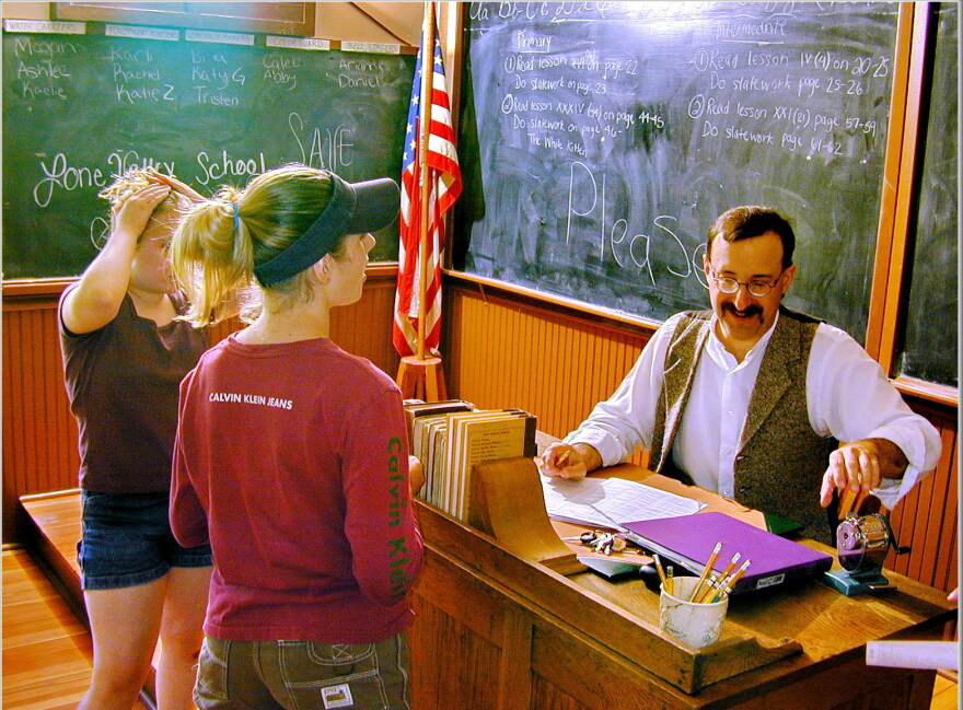 Randy Graubard sits at a wooden desk and reaches for an old pencil sharpener inside the historic Lone Valley School at Centennial Village. There's chalkboards behind him that read in part "Lone Valley School," "SAVE," and "PLEASE." There's two young women in front of him talking with him, one wearing a brown shirt and one wearing a red shirt. 