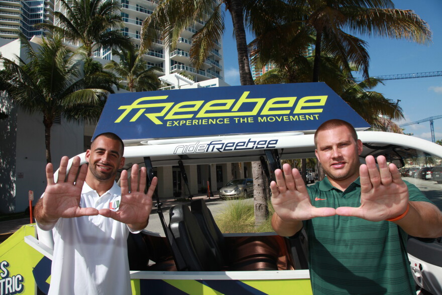 Two men holding their hands up in the shape of a U in front of a golf cart with the Freebee logo.
