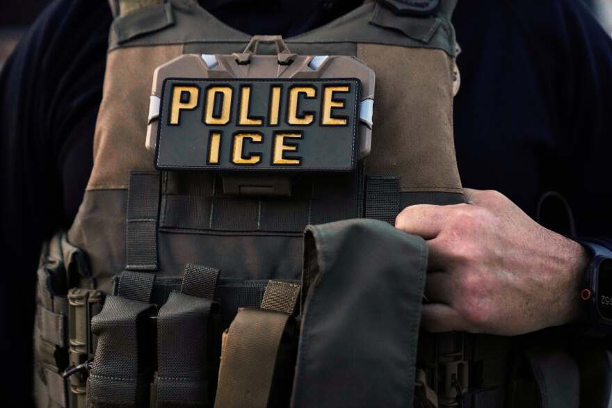 A light brown tactical vest is worn by an unseen man. One of his hands clasps the vest at the right side of the image. In the chest of the vest, a badge reads "Police, ICE."