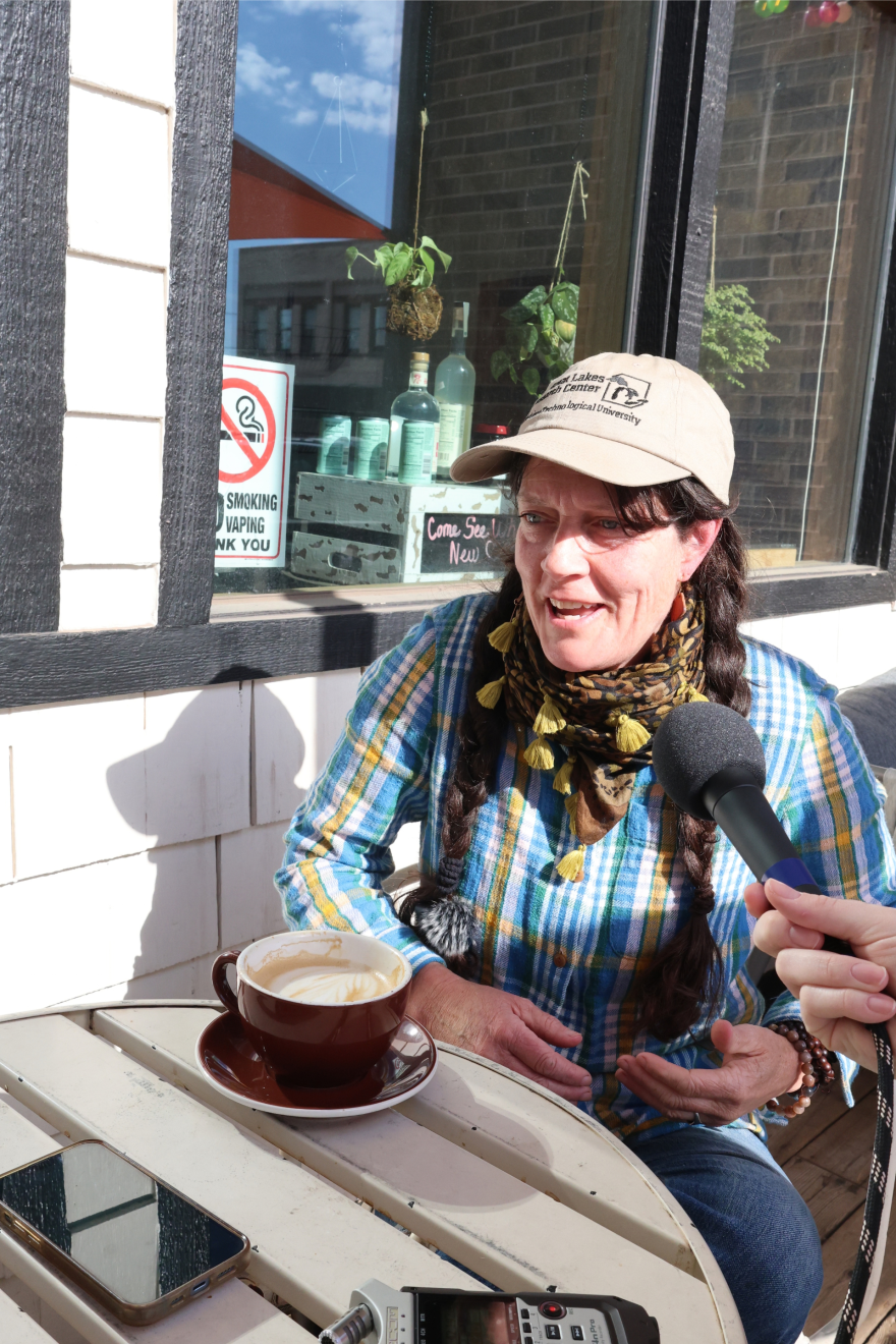 A white woman in a blue and yellow plaid shirt and a beige baseball cap speaks into a microphone. She is sitting at a white cafe table with a cup of coffee in front of her. 
