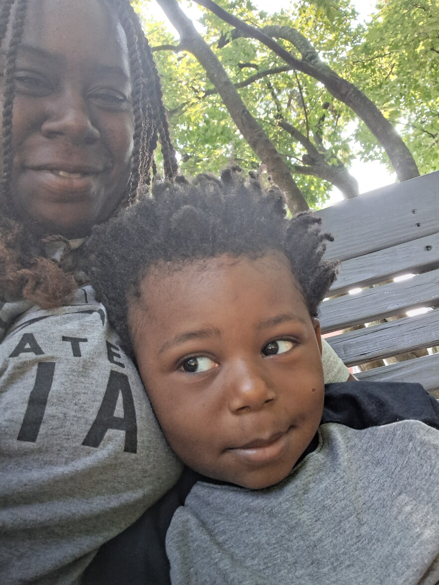 A woman and her son sit on a bench with trees in the background.