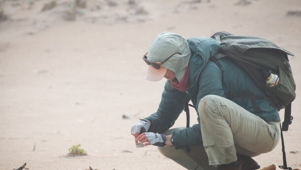 A woman crouches in the sand holding a small cup of water and a tiny egg.