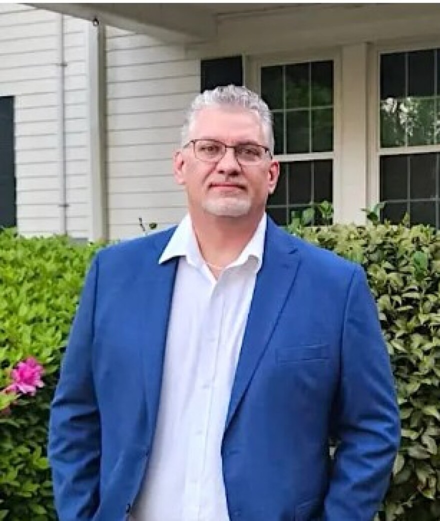Man with gray hair, glasses, wearing a blue sportcoat and hands in pockets, smiling into the camera