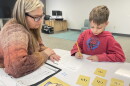 An adult in a sweater and glasses watches a child in a red hoodie complete a worksheet. 