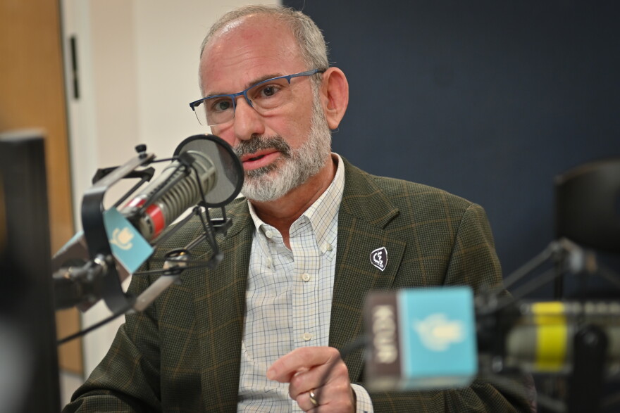 A man sits inside a radio studio. He is talking at a microphone and gesturing with his left hand.