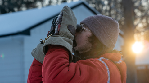A man in a red coat and large mittens peers through binoculars on a winter morning.